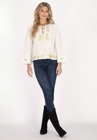 Weiße Bluse mit goldener Stickerei, Glockenärmel und Schnürung vorne. Kombiniert mit dunklen Skinny-Jeans und knielangen schwarzen Stiefeln.