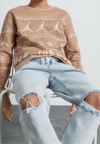 Felpa marrone chiaro con loghi Jordan bianchi e motivo a testo; abbinata a jeans denim azzurri strappati con bordi sfrangiati.