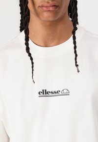 Sudadera de algodón blanca con un logo negro de "ellesse" con un acento curvado, cuello redondo y cabello trenzado visible en primer plano.