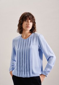 Blusa azzurro chiaro a maniche lunghe con plissè verticali sulla parte anteriore, scollo rotondo e polsini con bottoni. Tessuto liscio con una leggera lucentezza.