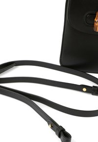 Sac à main en cuir noir avec des bretelles longues et fines et un fermoir de style bambou sur le rabat avant visible sur fond blanc.