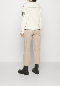 Witte trui met zwarte stiksels en logo op de mouw, gecombineerd met lichtbeige rechte broek en zwarte enkellaarsjes.