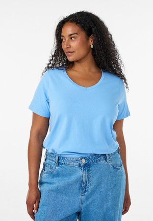 Femme aux cheveux bouclés et foncés portant un T-shirt bleu clair à col en V et un jean bleu taille haute, regardant vers sa gauche sur un fond blanc.
