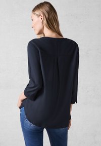 Navyblauw shirt met 3/4 mouwen, geplooid ontwerp aan de achterkant en een ronde zoom. De stof lijkt soepel en lichtgewicht. Gecombineerd met denim jeans.