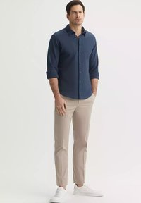 Granatowa koszula zapinana na guziki z podwiniętymi mankietami, zestawiona z jasnobeżowymi spodniami slim-fit i białymi trampkami, przedstawiona na neutralnym tle.