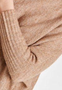 Měkký, světle hnědý svetr s ribovanými manžetami a texturovaným pleteným vzorem s jemnými variacemi v barvě a tloušťce.