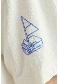Weißer Textilstoff mit blauem gesticktem Segelschiff-Design und dem Text "Sandieus" darunter. Bietet detaillierte Nähte und eine weiche Textur.