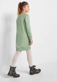Ragazza che cammina, indossando un vestito lungo verde chiaro con maniche lunghe, collant bianchi e stivali neri con lacci in pizzo floreale, di spalle su uno sfondo bianco.