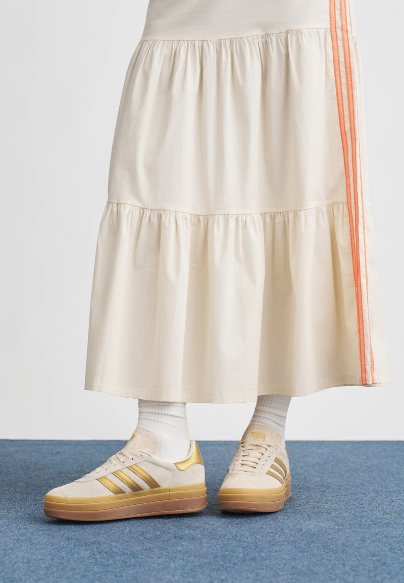 Beige sneakers with gold accents, textured suede and smooth rubber sole. Worn with an ivory tiered skirt featuring orange side stripes.