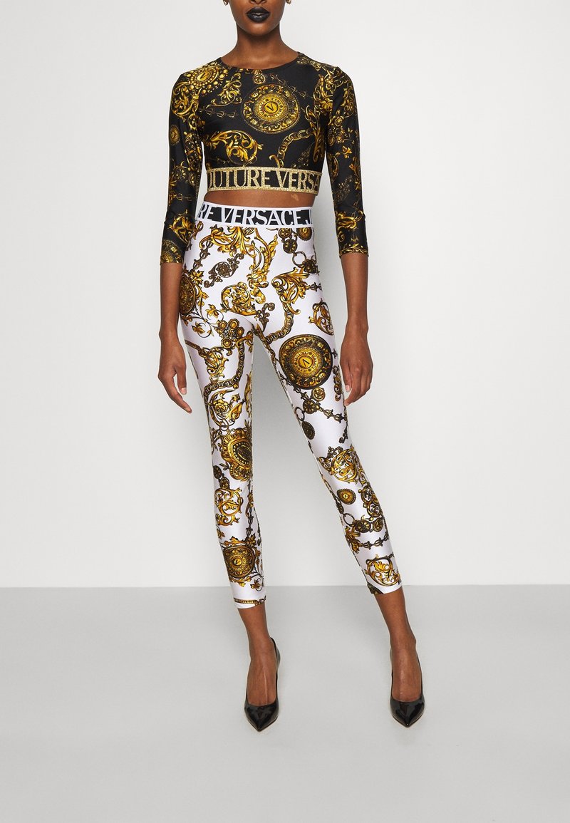 Zwart cropped top met lange mouwen en goudkleurig barokpatroon; witte leggings met gouden motieven. Zwarte hoge hakken, kenmerkende logoband rond de taille.