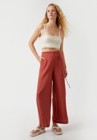 Pantalones anchos de lino rojos con cintura de cordón y bolsillos laterales, combinados con una blusa recortada de encaje blanca y sandalias neutras.