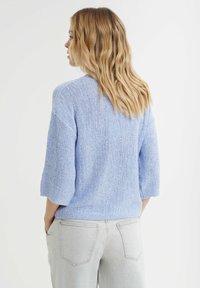 Lichtblauwe gebreide trui met een textuurafwerking, korte mouwen en een relaxte pasvorm, gecombineerd met lichtgrijze jeans.