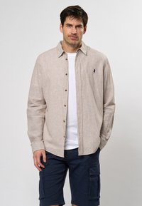 Camisa de botón color beige claro con un sutil patrón a rayas, mangas largas y un pequeño logo en azul marino. Combinada con pantalones cortos de cargo en azul marino.