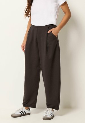 Femme portant un pantalon noir ample taille haute avec poches latérales, associé à un haut blanc à manches courtes et des baskets blanches avec des rayures noires.