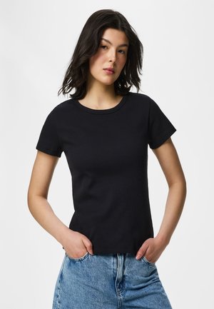 Jeune femme aux cheveux foncés mi-longs, portant un t-shirt noir à manches courtes et un jean bleu, mains dans les poches, expression neutre.