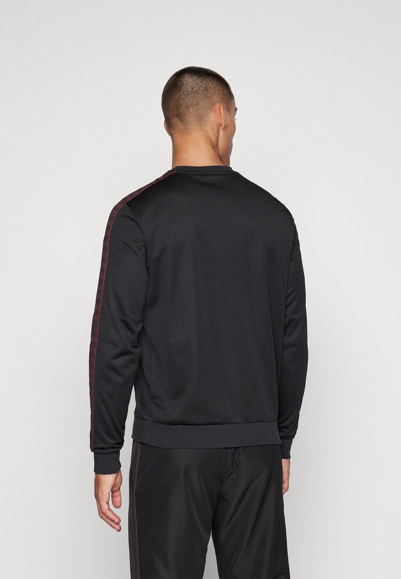 Sweatshirt de sport noir avec un col rond, présentant des rayures rouges le long des épaules. Fabriqué à partir d'un tissu lisse et extensible.