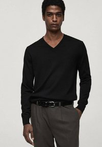 Schwarzer V-Ausschnitt-Pullover aus weichem Material, mit langen Ärmeln, kombiniert mit maßgeschneiderten Hosen und einem schwarzen Gürtel, vor einem hellen Hintergrund.