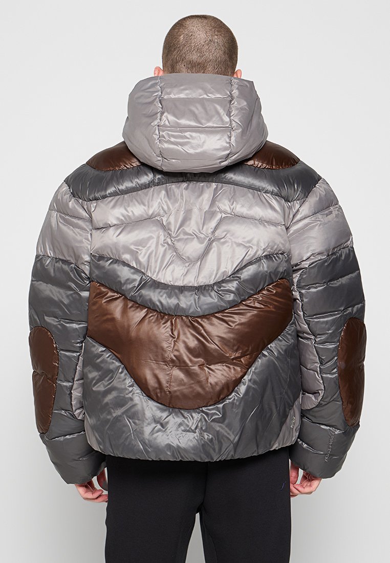 Chaqueta tipo pluma que presenta una mezcla de telas grises y marrones, con capucha y patrones abstractos acolchados con acentos redondeados.