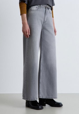 Personne portant un jean large gris clair, des bottines noires, un t-shirt à manches longues moutarde sous un haut gris foncé, debout sur un sol clair.