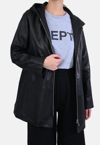 Chaqueta de cuero negra con cremallera frontal, capucha y dos grandes bolsillos delanteros. Llevada sobre una camisa gris con letras negras llamativas.