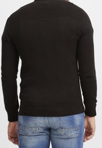 Mężczyzna w czarnej bluzie z długim rękawem i niebieskich dżinsach, przedstawiony z tyłu z rękami swobodnie opadającymi wzdłuż ciała.