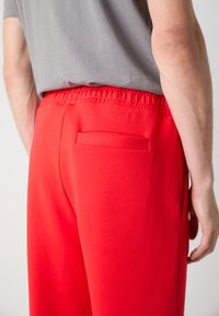 Rode joggingsbroek met een elastische tailleband, voorzien van een achterzak en een gladde stofafwerking. Draagt een lichtgrijze shirt.