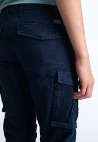 Námořnické cargo kalhoty z bavlny s několika kapsami, nášivkou a hladkou texturou. Fit je zobrazen zezadu s rukou na boku.