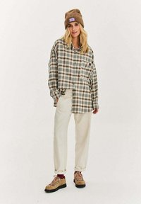 Kobieta w beżowej czapce typu beanie, oversize'owej koszuli w kratę, kremowych spodniach i jasnobrązowych butach turystycznych z różowymi sznurówkami na jednolitym tle.