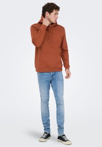 Bruine hoodie met een voorzak, capuchon met trekkoord en geribbelde zoom. Gecombineerd met lichtblauwe jeans en zwarte sneakers.
