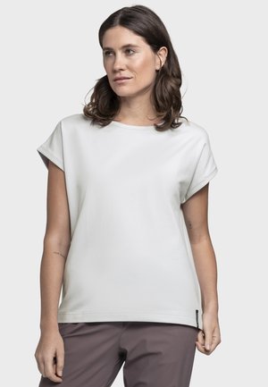 Frau mit schulterlangem braunem Haar, die ein weißes Kurzarmshirt und braune Hosen trägt, vor einem schlichten hellen Hintergrund stehend.