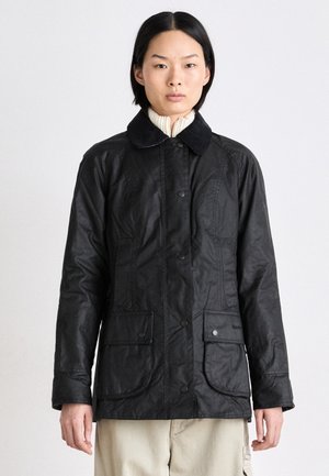 Veste en tissu ciré noir avec un col en velours côtelé, des boutons-pression et deux poches avant. Présente une coupe ajustée et des détails de couture visibles.