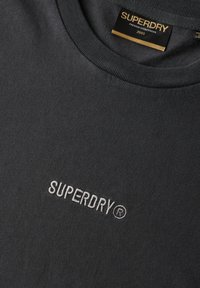 Camiseta de cuello redondo en gris oscuro hecha de material acanalado, con el logotipo bordado "SUPERDRY" en gris claro en el frente.