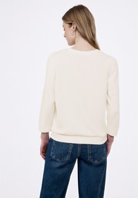 Kremowa bluzka z długim rękawem, okrągłym dekoltem, miękką fakturą i mankietami z prążkowanej dzianiny, zestawiona z niebieskimi dżinsami.
