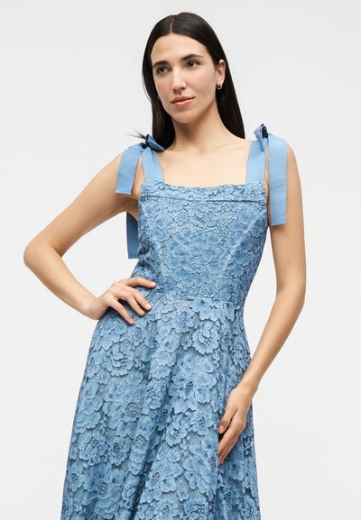 Γυναίκα με μακριά σκοτεινά μαλλιά φοράει μπλε αμάνικο δαντελωτό φόρεμα με λουλούδια και κορδόνια στους ώμους, ποζάροντας με το ένα χέρι στη μέση και το άλλο αγγίζοντας τη φούστα.