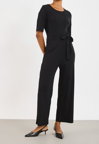 Černý žebrovaný overal s krátkými rukávy, se staženým pasem svázaným látkovým pásem, doplněný černými lodičkami s puntíky a otevřenou patou.