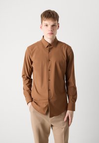Camicia button-up marrone in tessuto liscio, con maniche lunghe e colletto classico. Abbinata a pantaloni beige. I bottoni sono dello stesso colore della camicia.
