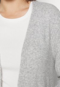 Cardigan grigio chiaro realizzato in materiale morbido, con apertura frontale, dettagli a coste lungo i bordi e vestibilità rilassata, indossato sopra una maglietta bianca.