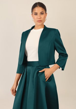 Türkise, taillierte Jacke mit Dreiviertelärmeln über einem weißen Oberteil. Der Rock ist plissiert und aus einem ähnlichen türkisen Stoff gefertigt, um einen abgestimmten Look zu erzielen.