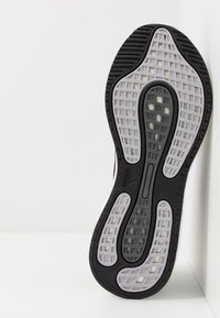 Semelle de chaussure de course Adidas noire et grise avec motif de bande de roulement en forme de grille, positionnée verticalement contre un mur blanc et un sol blanc.