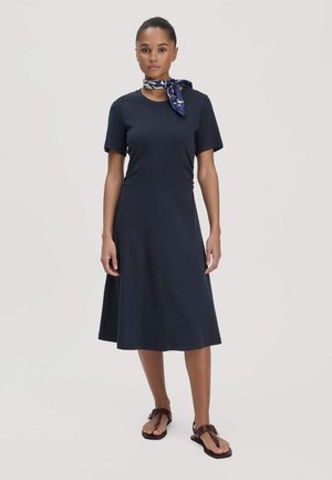 Frau trägt ein marineblaues Midi-Kleid mit kurzen Ärmeln, braune Sandalen mit Schnalle und ein blau gemustertes Halstuch, steht vor einem schlichten Hintergrund.