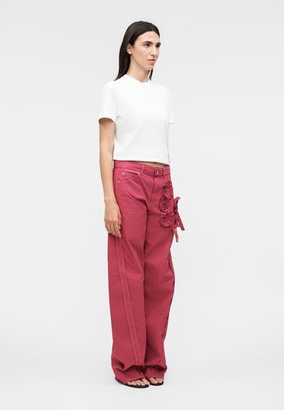 Γυναίκα στέκεται φορώντας λευκό crop top με κοντά μανίκια και κόκκινο παντελόνι ψηλόμεσο με φαρδιά πόδια και διακοσμητικά υφασμάτινα λουλούδια στο ισχίο.