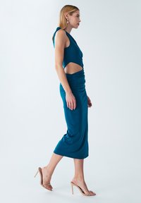 Teal-farbenes, figurbetontes Kleid mit einem Ausschnitt in der Taille und Raffung seitlich, kombiniert mit beigen, geschnürten High Heels.