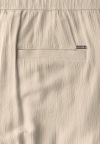 Beige textured fabric with crinkled appearance. Features a rectangular pocket and a silver "Cecil" tag. Smooth, lightweight material.