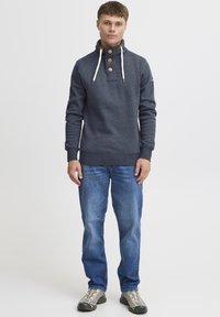 Navy sweatshirt met hoge kraag, drie bruine knopen en crèmekleurige koorden, gecombineerd met blauwe denim jeans en grijze schoenen.