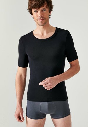 Black short-sleeve shirt made of smooth fabric, featuring a round neckline. Model wearing gray shorts, standing against a neutral background.