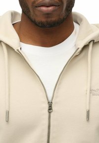 Felpa beige con zip, dal tessuto morbido, con zip argentata, cordini e una t-shirt bianca sotto. Dettaglio logo sottile sul tessuto.