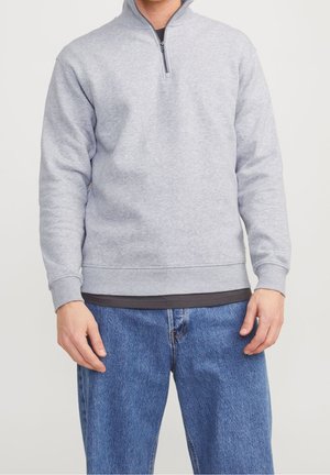 Hombre con sudadera gris claro de media cremallera sobre camisa oscura y vaqueros azules, de pie frente a un fondo claro y liso.