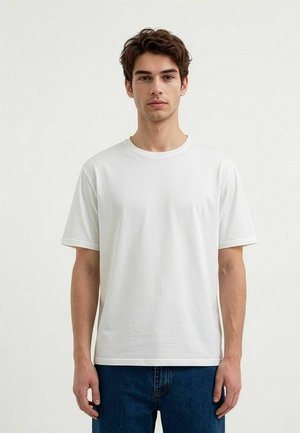 Jonge man met kort bruin haar die een effen wit T-shirt met korte mouwen en blauwe spijkerbroek draagt, staand tegen een witte achtergrond.