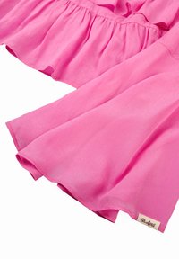 Vaaleanpunainen röyhelöbluse kevyestä kankaasta, jossa on kerroksellinen muotoilu, liehuvat hihat ja pieni etiketti helman kohdalla.