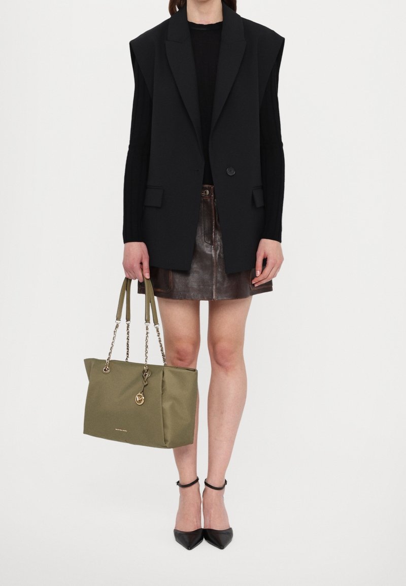 Femme portant un blazer noir sans manches, un haut noir à manches longues, une jupe courte en cuir marron, des talons noirs à bride à la cheville, tenant un sac à main vert olive.